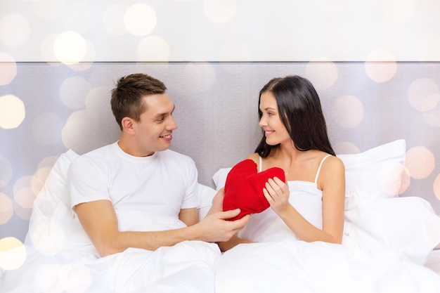 love, relationships, holidays and people concept - smiling couple in bed with red heart-shaped pillow over lights background