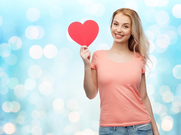 love, romance, charity, valentines day and people concept - smiling young woman or teenage girl with blank red heart shape over blue holidays lights background