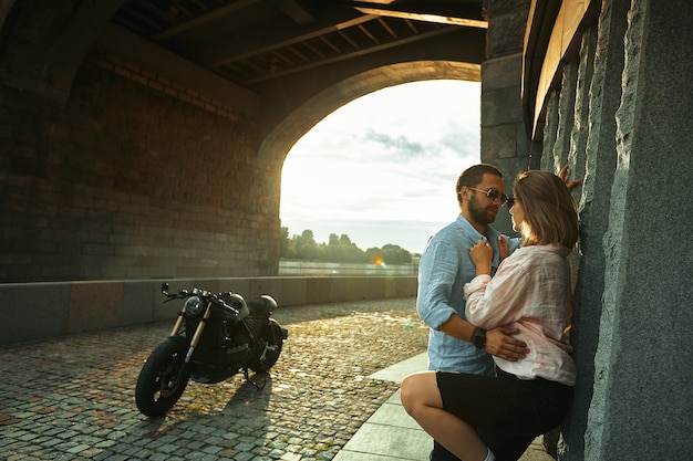 Love and romantic concept. Couple in love with sunset stand by the wall kiss and hug under the bridge next to motorcycle. Man with beard hugging womans, tenderness.