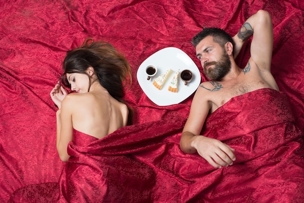 Love and sex concept. Couple in love on burgundy sheets. Man and woman with half covered bodies lie in bedroom. Man with beard and serious face has romantic breakfast with dreamy lady in bed, top view