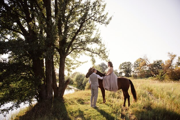 Love story and wedding near the river in a field at sunset with brown horse. The bride in airy dress is the color of dusty rose. Beige dress with sparkles. bride and bridegroom embrace and kiss.