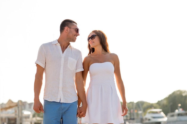 love, travel, tourism, sailing, people and friendship concept - smiling couple wearing sunglasses walking at harbor