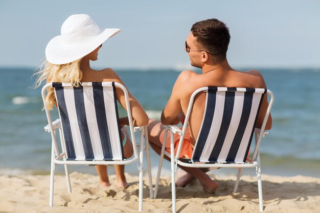 愛、旅行、観光、夏、人々 のコンセプト - 椅子に座って後ろからビーチで日光浴をする水着姿でバカンスに笑顔のカップル
