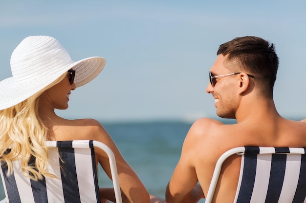 愛、旅行、観光、夏、人々 のコンセプト - 椅子に座って後ろからビーチで日光浴をする水着姿でバカンスに笑顔のカップル