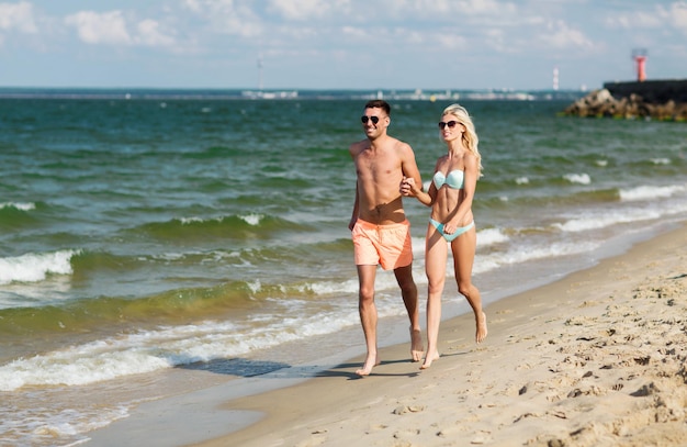 love, travel, tourism, summer and people concept - smiling couple on vacation in swimwear and sunglasses holding hands and running on beach