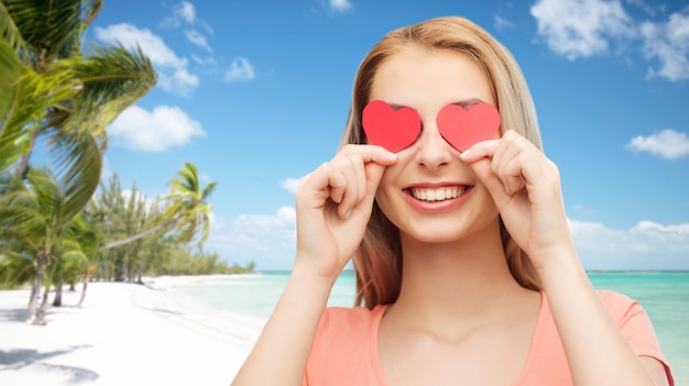любовь, путешествия, туризм, день святого валентина и концепция людей - улыбающаяся молодая женщина или девочка-подросток с красными сердечками на глазах над экзотическим тропическим пляжем на фоне пальм