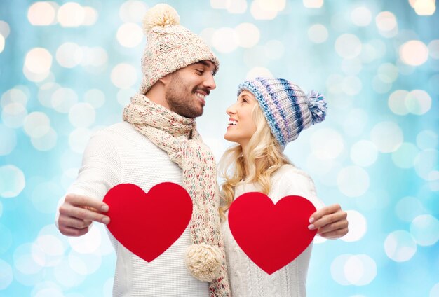 love, valentines day, couple, christmas and people concept - smiling man and woman in winter hats and scarf holding red paper heart shapes over blue holidays lights background