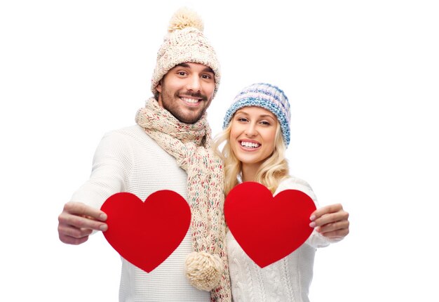 love, valentines day, couple, christmas and people concept - smiling man and woman in winter hats and scarf holding red paper heart shapes