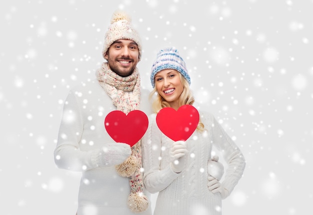 love, valentines day, couple, christmas and people concept - smiling man and woman in winter hats and scarf holding red paper heart shapes