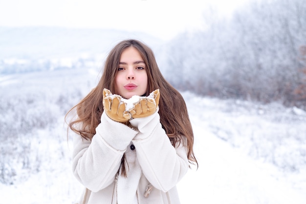 冬が大好きです。赤い唇を吹く幸せなクールな女の子は、雪の背景の上に白いセーターを着てエアキスをします。