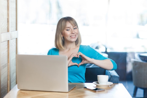 Love you! Portrait of romantic happy young girl with blonde hair in blue blouse are sitting in cafe and looking at display with a heart shape with fingers through a webcam online video call. Indoor