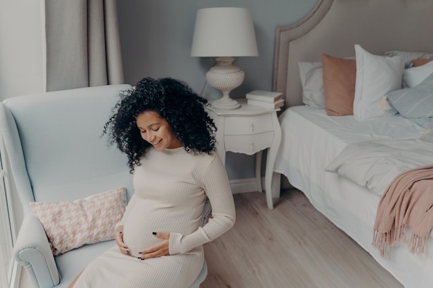 침실에 앉아 행복한 임신을 즐기는 사랑스러운 아프리카계 미국인
