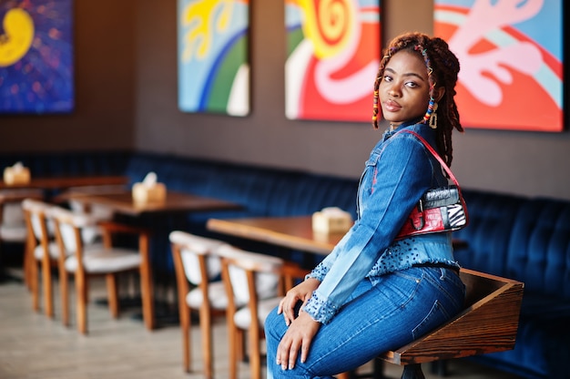 Прекрасная афро-американская женщина с дредами в синей стильной джинсовой куртке в кафе. Красивая прохладная модная черная маленькая девочка в помещении.