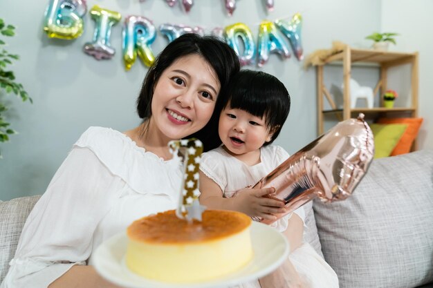 사랑스러운 아시아 아기 아이가 호기심을 가지고 케이크를 보고 있는 동안 그녀의 어머니는 생일에 가까이 포즈를 취하고 카메라를 향해 미소를 짓고 있습니다