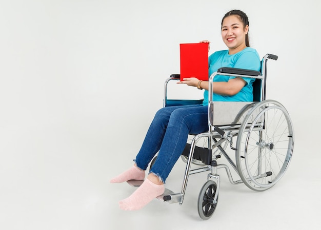 사랑스러운 아시아 장애 여성은 신체적 부상에서 재활을 한 후 마비 병원의 의료 휠체어에 앉아 있는 동안 재활 클리닉의 빨간색 의료 파일을 보여주기 위해 행복하고 미소를 지었습니다.