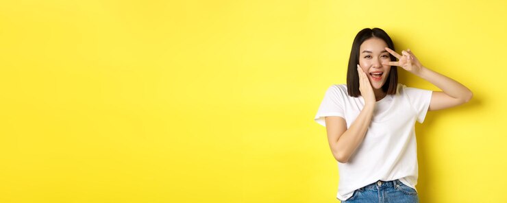 Photo 白い t シャツを着た素敵なアジアの女の子が頬に手を当てて立っている目でピースサインを示す