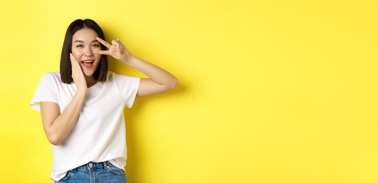 Photo 白い t シャツを着た素敵なアジアの女の子が頬に手を当てて立っている目でピースサインを示す