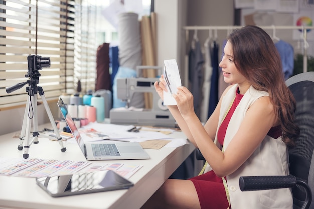 A lovely Asian woman fashion designer sits at her workstation in her studio, stares at the webcam, and speaks through video call.