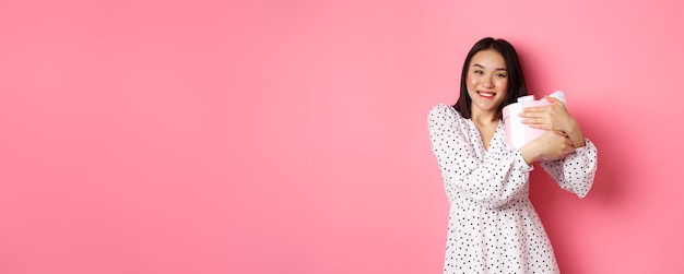 Lovely asian woman hugging her gift and smiling thankful receive valentines day present standing ove