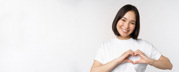 Lovely asian woman smiling showing heart sign express tenderness and affection standing over white b