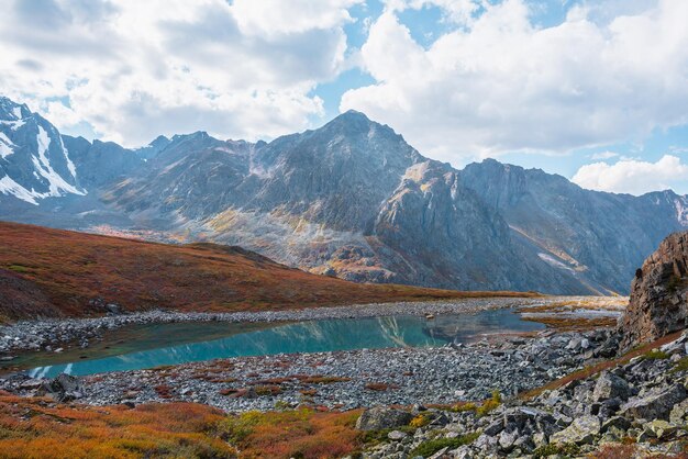 Lovely autumn landscape with turquoise mountain lake against high mountain top in sunlight under clouds in blue sky Sunlit beautiful small alpine lake and large mountain range Vivid autumn colors