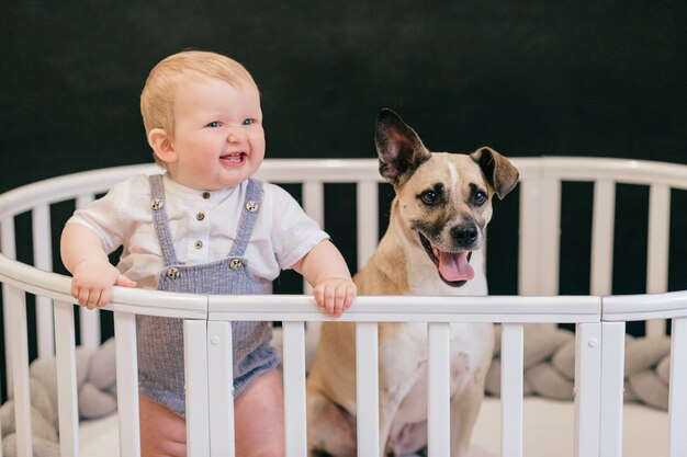 Lovely baby boy with dog standing together in crib over black background