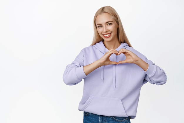 Lovely blond woman showing heart sign and smiling, wearing hoodie with jeans, standing on white