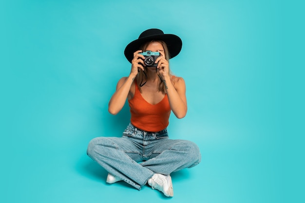 Lovely blond woman in  stylish summer outfit holding retro camera , sitting on floor in studio on blue wall. Vacation mood.