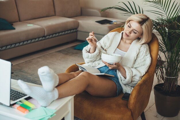 Photo lovely blonde woman is sitting in armchair and watching something on a laptop while eating cereals