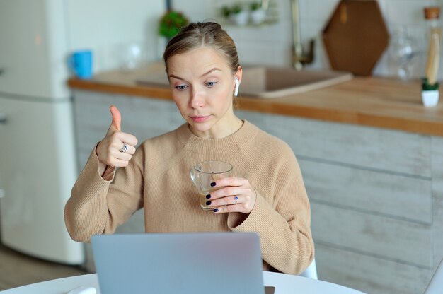 Lovely blonde woman working with her laptop while sitting in the kitchen in her apartment