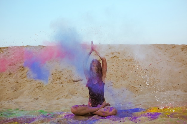 Foto bella donna bruna con i capelli lunghi che gioca con la vernice holi, seduta sulla sabbia