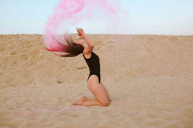 사진 핑크 홀리 페인트 구름과 함께 포즈를 취하는 긴 머리를 가진 사랑스러운 갈색 머리 여자, 모래에 앉아