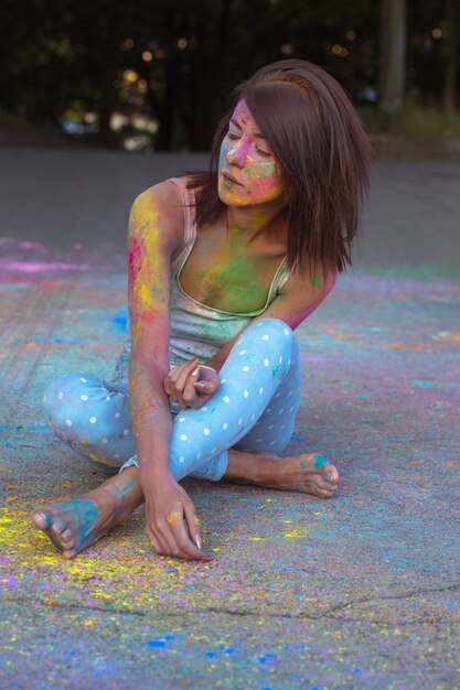 Lovely brunette woman with short hair covered Holi powder, sitting on the asphalt