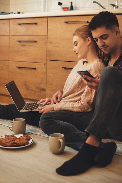 Bella coppia caucasica seduti in cucina sul pavimento navigando in un laptop e telefono mentre si consuma una colazione