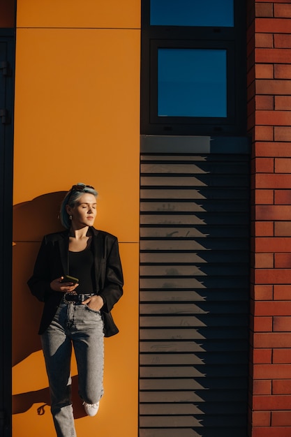 Lovely caucasian girl with blue hair posing on a yellow wall outside wearing eyeglasses and holding a phone