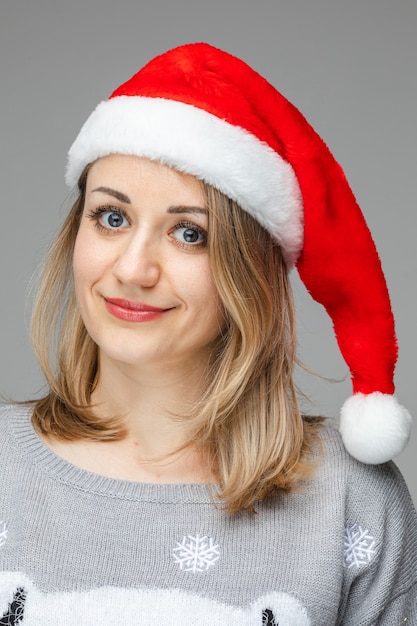 Lovely Caucasian woman with blonde hair and red lips in red Santa hat and grey sweater smiling