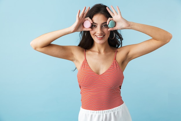 Lovely cheerful girl wearing summer clothes standing isolated over blue, eating tasty macaroons