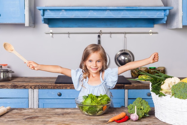 Lovely child girl joyfully raising her hands holding wooden spoon and smiling looking at camera happy to cook salad of fresh green vegetables preparing healthy breakfast vegetarian food vitamins