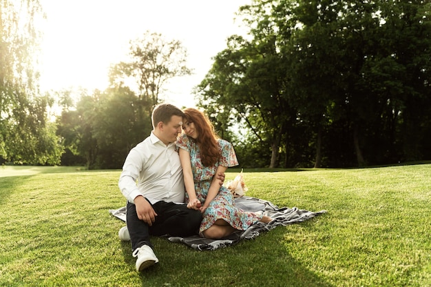 愛する素敵なカップルが公園でピクニックをし、ベッドカバーに花と食べ物が入った籐のバスケットを持っていました。幸せな恋人たちはピクニックで笑って食べます。ロマンチックなデート