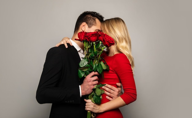 A lovely couple in smart outfits are hiding behind a beautiful bouquet of red roses while kissing and hugging.