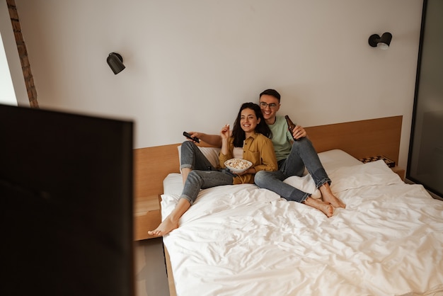 Прекрасная пара с тарелкой поп-конра во время просмотра фильма по телевизору, сидя на кровати. счастливая молодая семья переехала в новую квартиру.