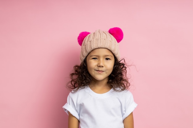 Lovely cunning little girl wearing white t shirt, knitted beige hat, looking sly and tricky, grinning mysteriously, having interesting plan or idea, standing. Facial expressions.