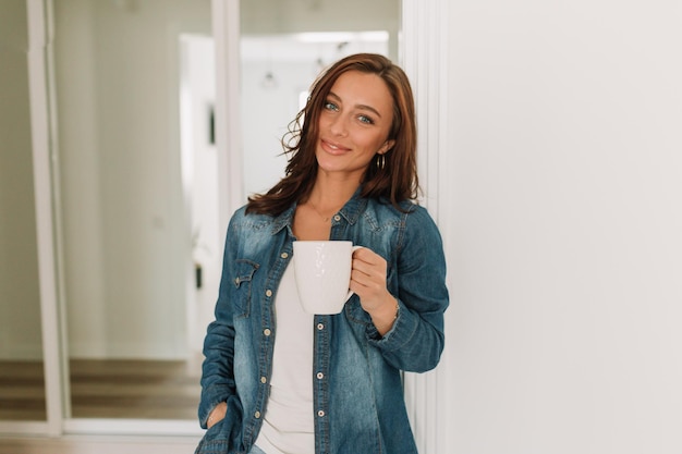 Bella donna carina con capelli ondulati scuri che indossa una camicia di jeans in piedi sullo sfondo del muro bianco con tè e guardando la fotocamera