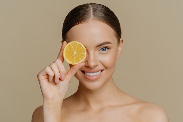 Lovely dark haired European woman smiles gently covers eye with slice of juicy lemon poses half naked