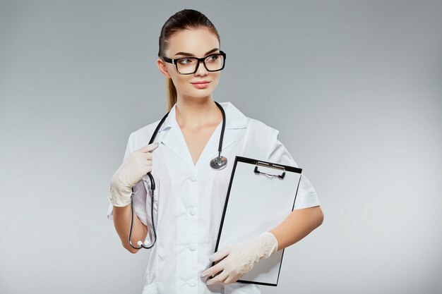 Lovely doctor with brown hair and nude make up wearing white medical uniform, glasses, stethoscopes and white gloves at gray studio background and holding notes.