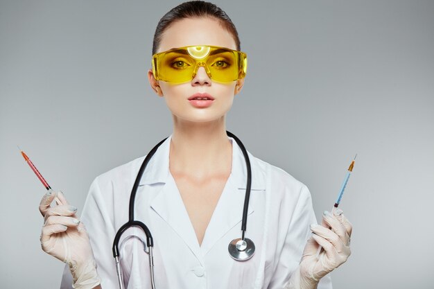 Lovely doctor with brown hair and nude make up wearing white medical uniform, glasses, stethoscopes and white gloves at gray  wall and holding syringes, portrait.