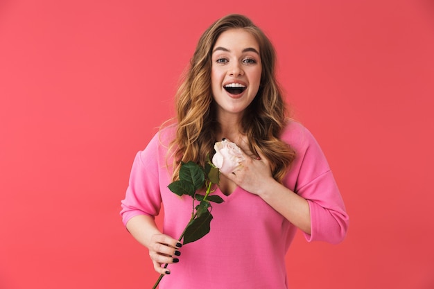 Lovely excited young blonde woman standing isolated over pink wall, holding white rose