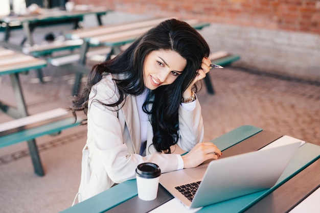 Bella donna d'affari con capelli lunghi scuri che tiene la penna in mano seduta all'aperto utilizzando il computer portatile per il suo lavoro a bere caffè cercando di realizzare un progetto adorabile donna bruna che lavora