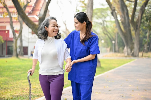 Lovely female caregiver assisting and supporting an old lady to walk with a walking stick