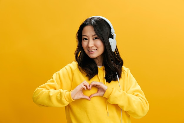 Lovely friendly cheerful asian student young woman in yellow\
hoodie sweatshirt show heart shaped sign posing isolated on over\
orange studio background cool offer perfect playlist sound app\
concept